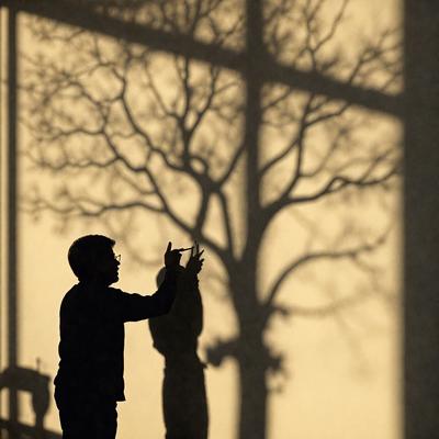 The primary subject of the image is a man standing in front of a window, looking at his reflection. The setting is indoors, with the man standing in front of a window. The visual style appears to be a photo, capturing the man's reflection in the window. The colors in the image are predominantly dark, with the man's reflection being the main focus. The overall atmosphere of the image is somewhat mysterious and introspective, as the man seems to be contemplating his reflection.